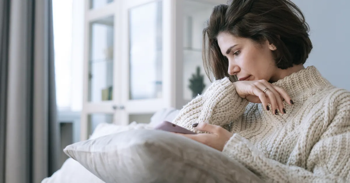 A woman in a knitted sweater sits pensively on a couch at home, using a smartphone.