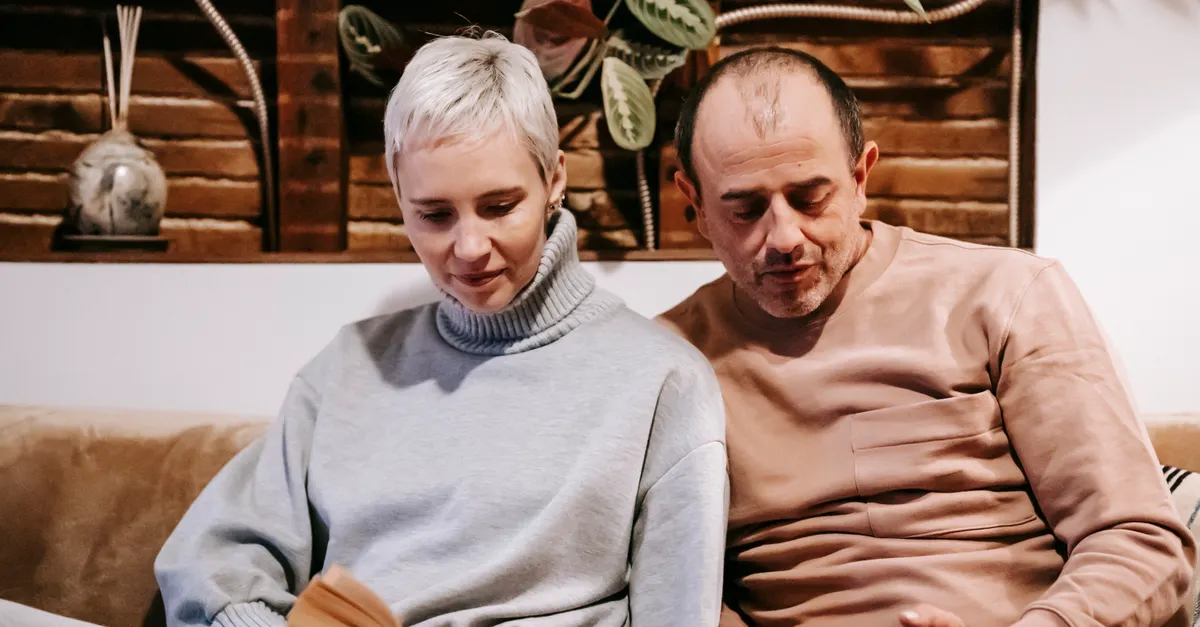 A middle-aged couple sitting on a sofa, one reading a book and the other using a tablet.