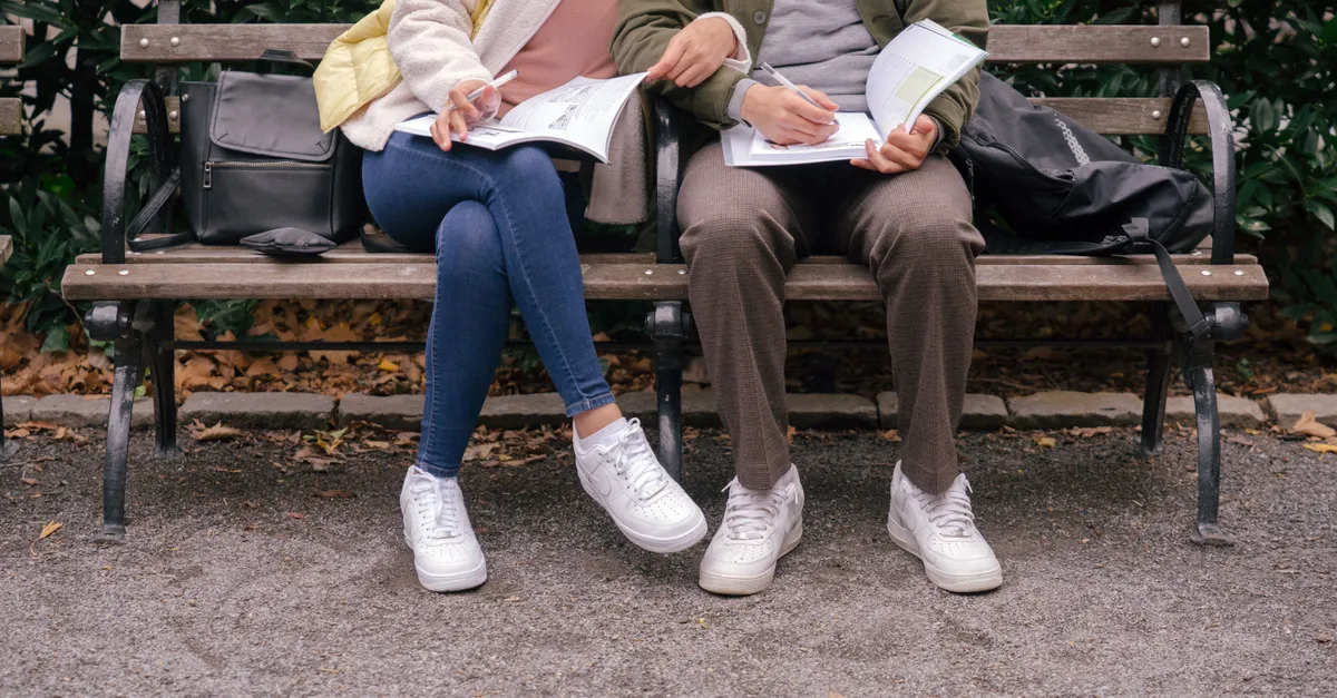 Couple in casual clothes on a park bench, doing homework in workbooks.