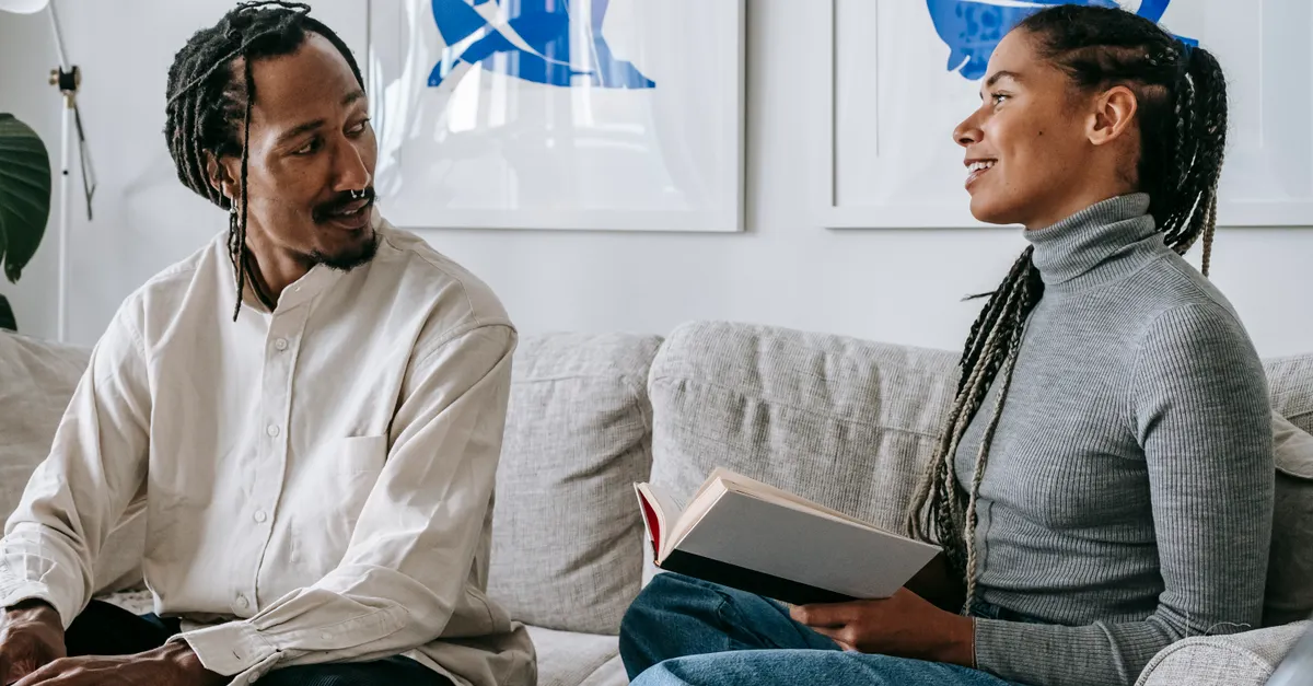 Woman with braids on a sofa holding a book, talking with her boyfriend who works on a laptop