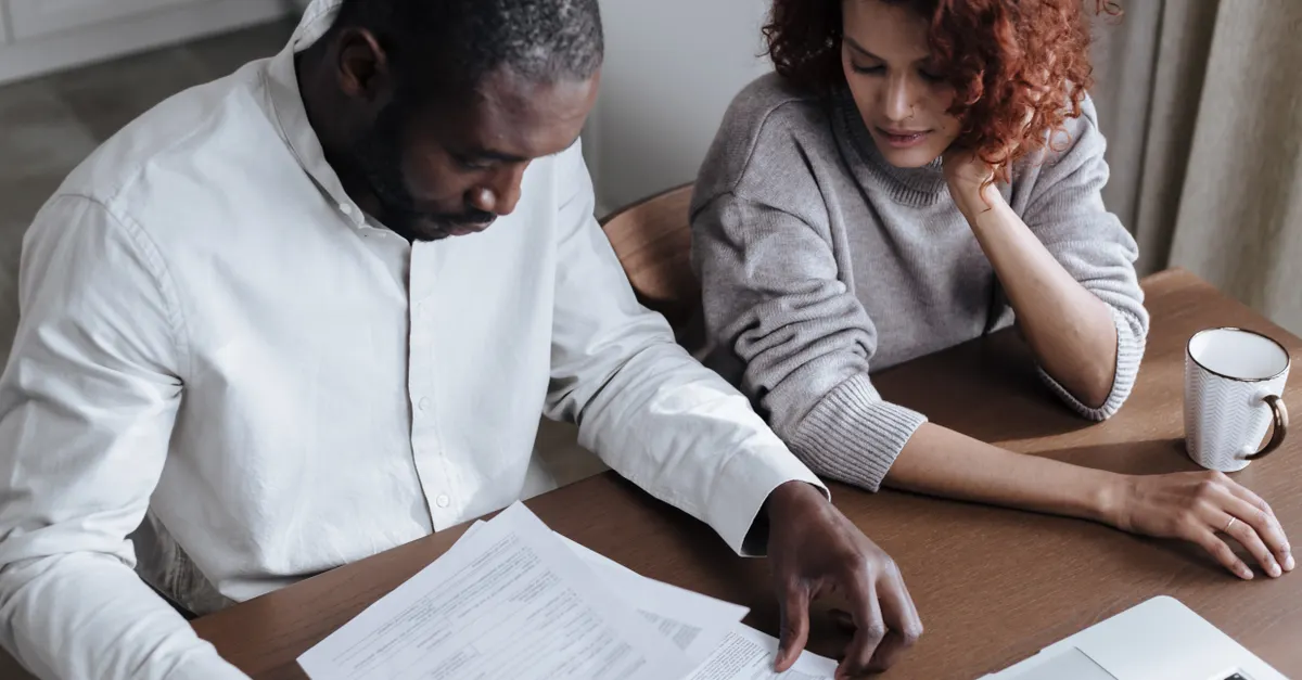 A couple sitting together at a home desk reviewing documents with a laptop.