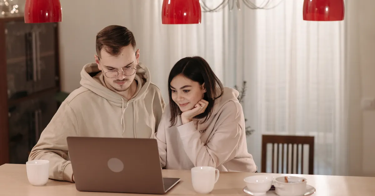 A couple in casual hoodies using a laptop at home, smiling and relaxed.