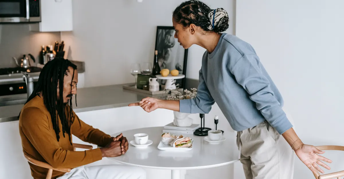 Young black woman blaming ethnic depressed boyfriend sitting at table in living room at home
