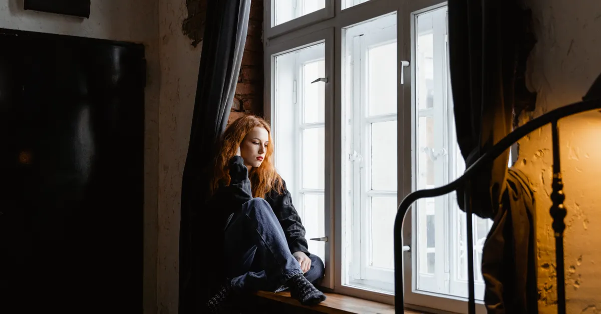 A thoughtful woman sits by a window in a cozy room, deep in contemplation.