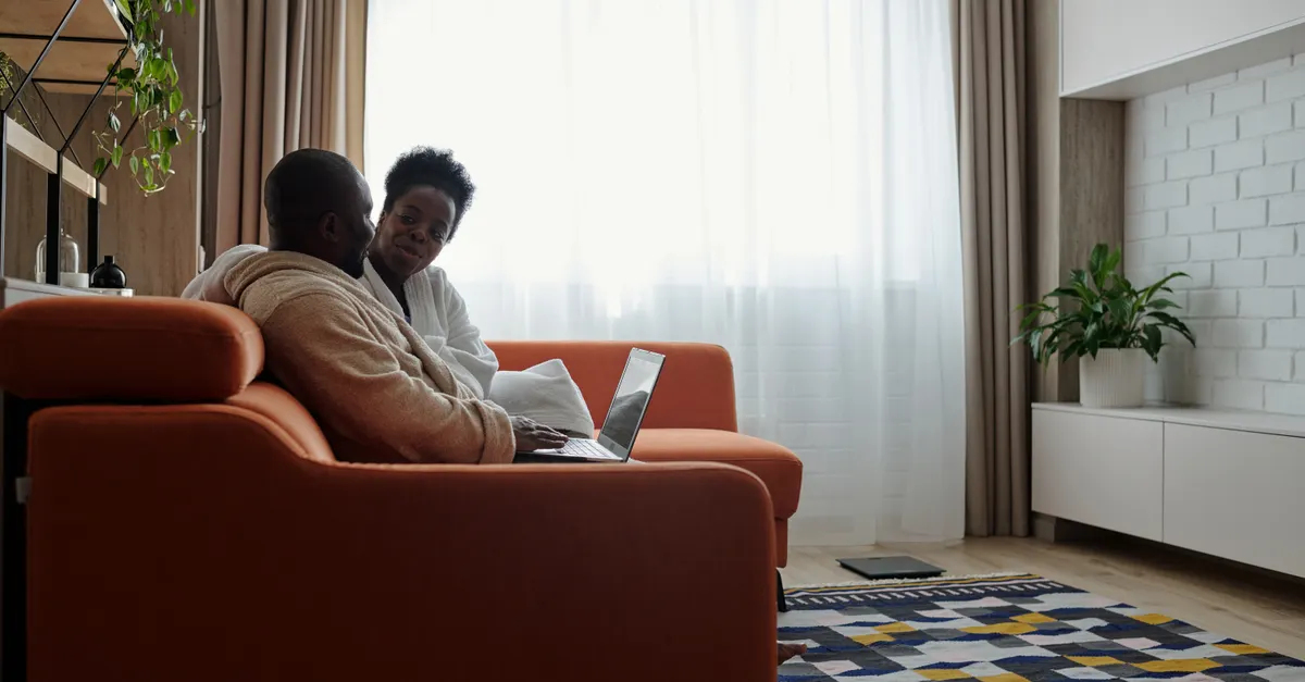 A couple sits on a sofa in a living room, talking while using a laptop.