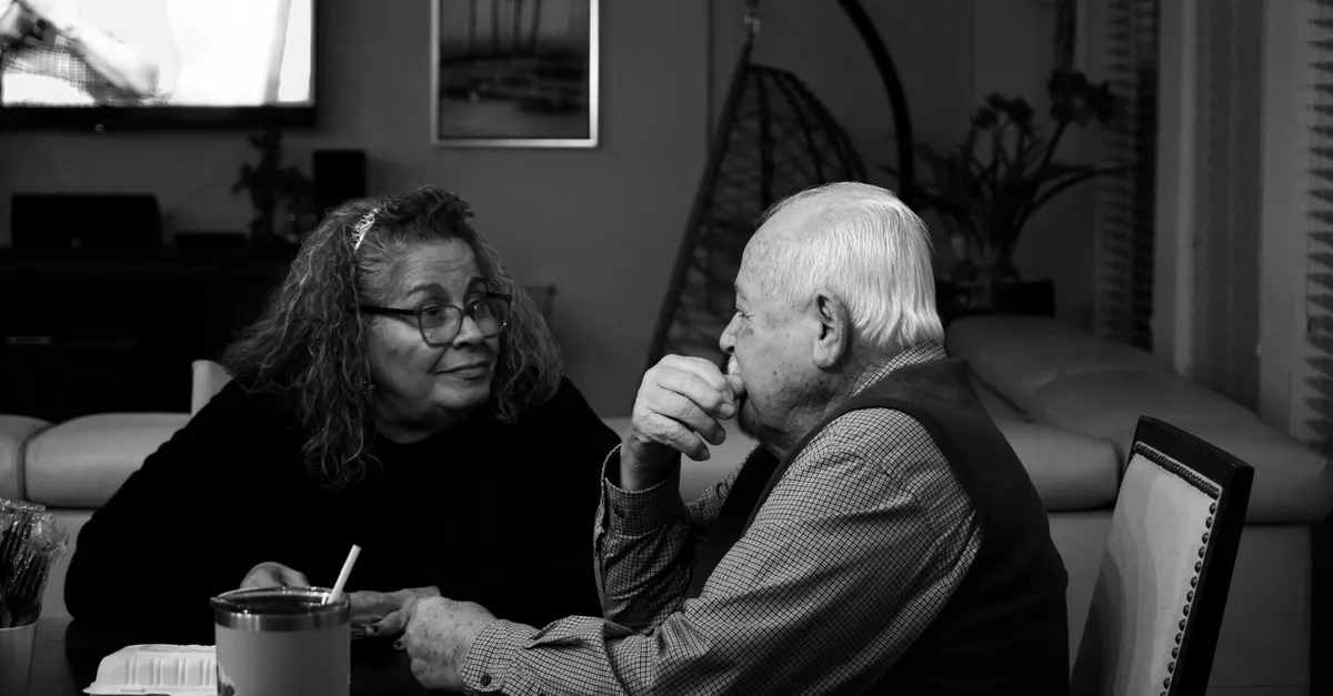 An elderly couple sitting across from each other in quiet conversation at an indoor dining table.