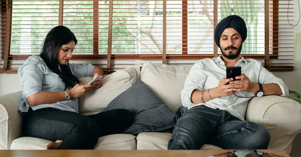 A serious bearded man in a turban and a woman in casual clothes sit on a couch, concentrating on their smartphones.
