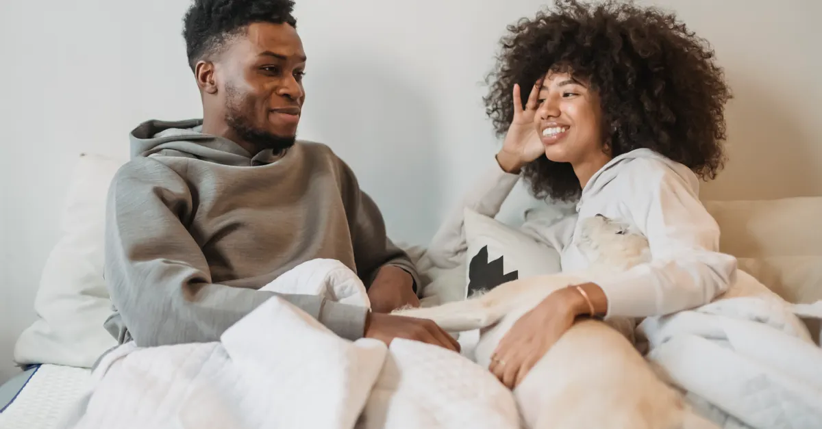 Happy African American couple in casual clothes lying on bed with cute dog and chatting.