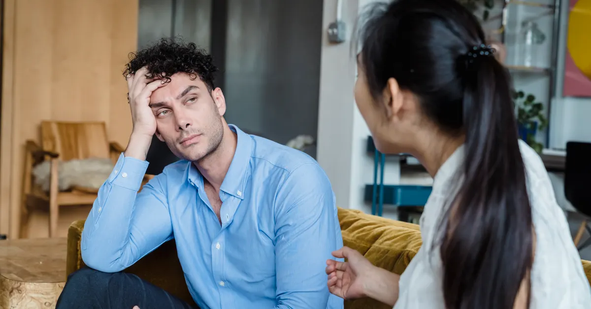 A tired man in a blue shirt listens while a woman talks indoors in a modern living room.