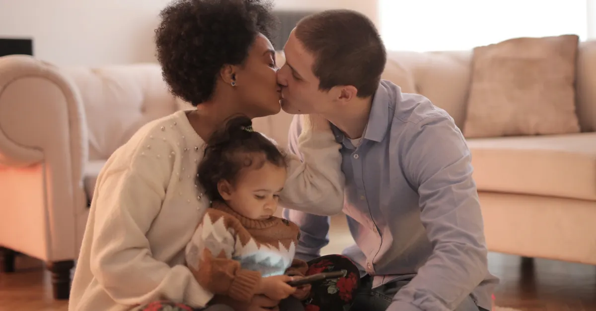 A loving couple kisses while sitting with their child in a warm living room setting.