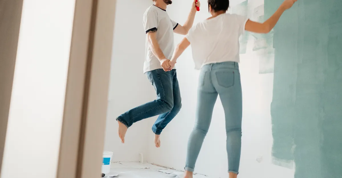 A couple in aprons painting a wall together, holding hands while laughing.