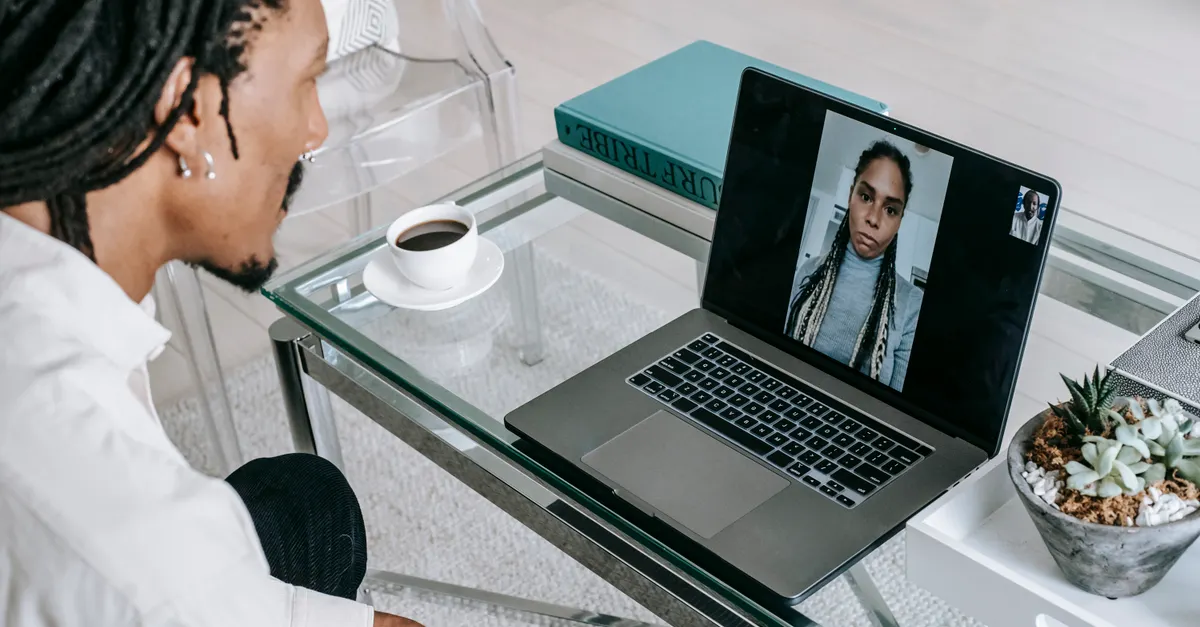 Man with dreadlocks video calling on laptop at a glass table with coffee and books.