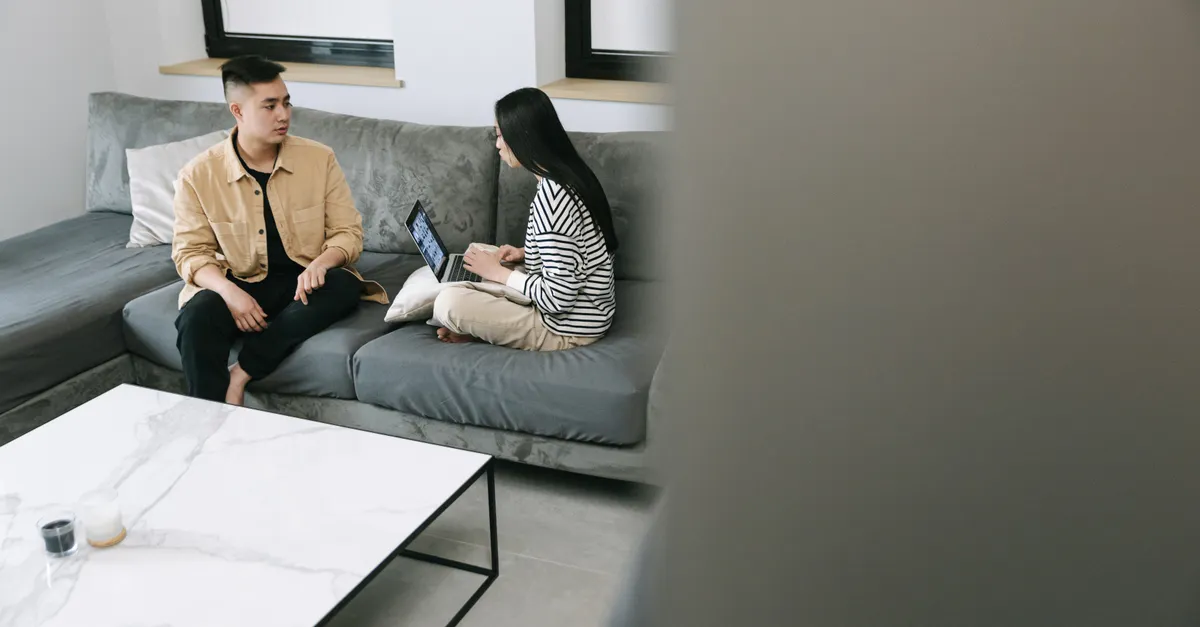 A couple sits on a sofa using a laptop, discussing something.