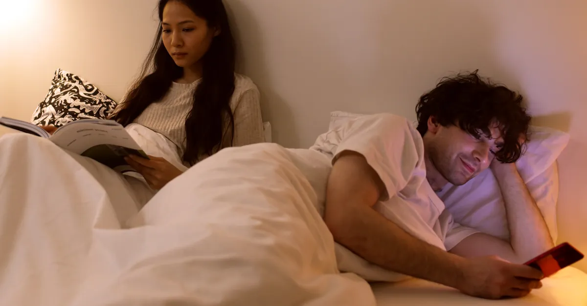 A couple in bed, one reading while the other uses a phone.