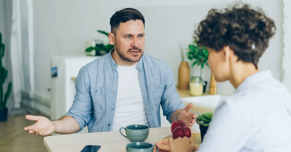 Two adults sitting at a kitchen table with coffee cups, talking together