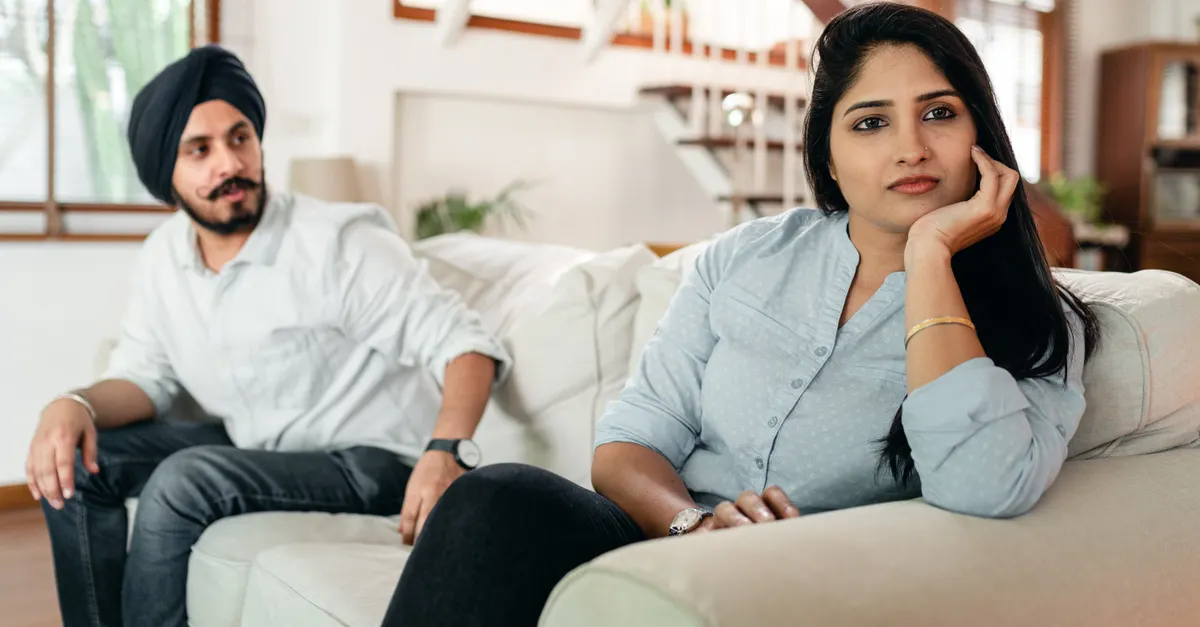 Offended Indian woman on sofa looking sadly away, bearded husband trying to make peace after quarrel.