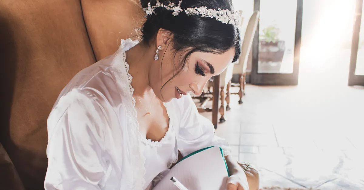 Bride in a wedding robe writing notes in a notebook during her wedding morning preparation.