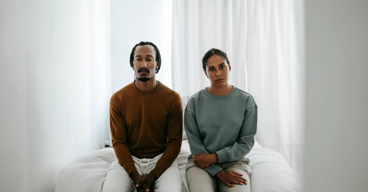 Young displeased African American couple in casual clothes sitting on bed and looking at camera during dispute