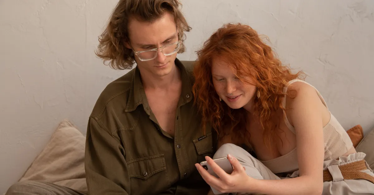 Calm couple in casual clothes resting together, browsing a smartphone in a light room.