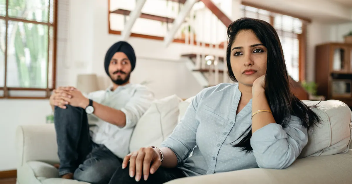 An Indian woman sitting on a sofa looking away from her husband who sits at the other end, appearing upset.