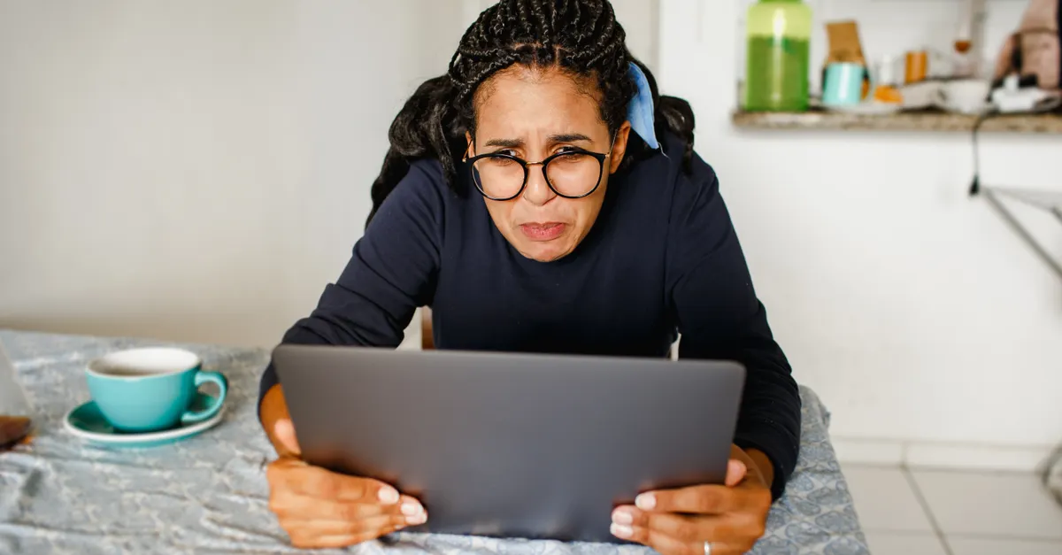 A woman wearing glasses looks stressed while using her laptop in a cozy kitchen