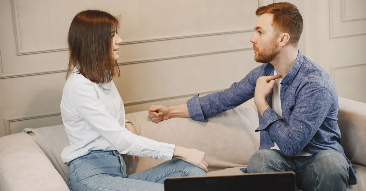 A young couple sitting on a beige couch indoors, engaged in conversation.