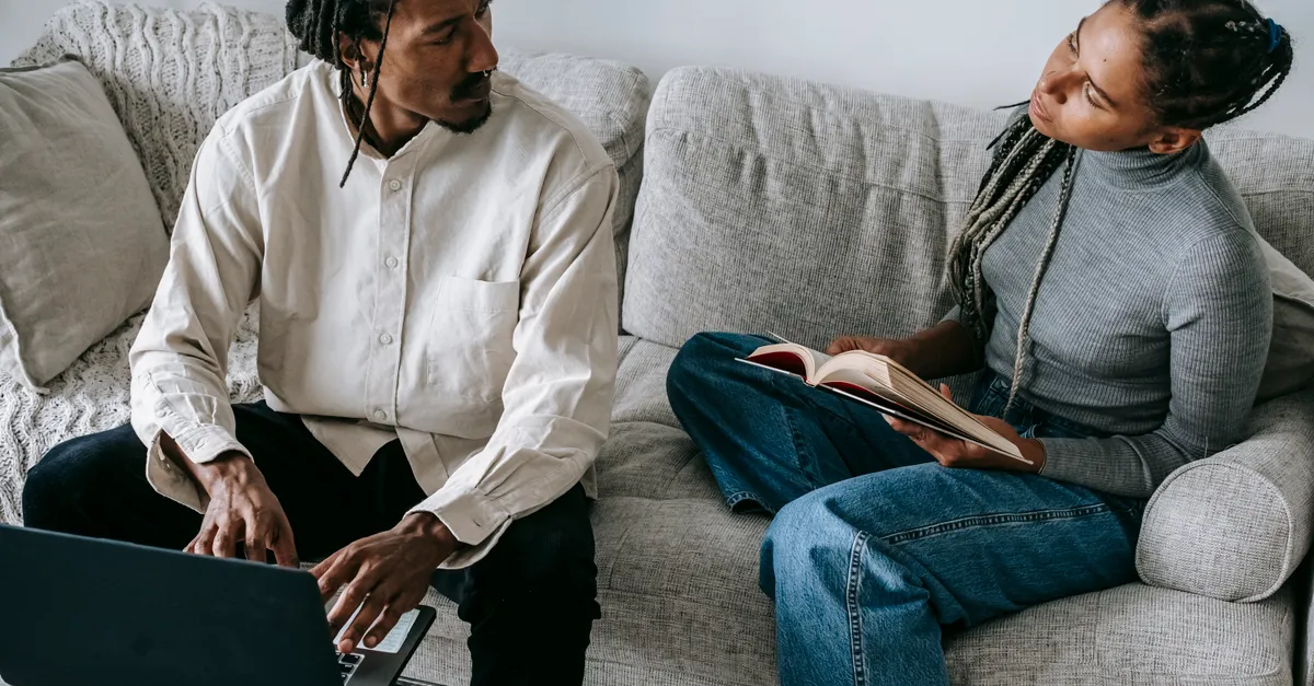 Serious couple sitting on a sofa with a book and laptop, discussing their relationship in a cozy living room.