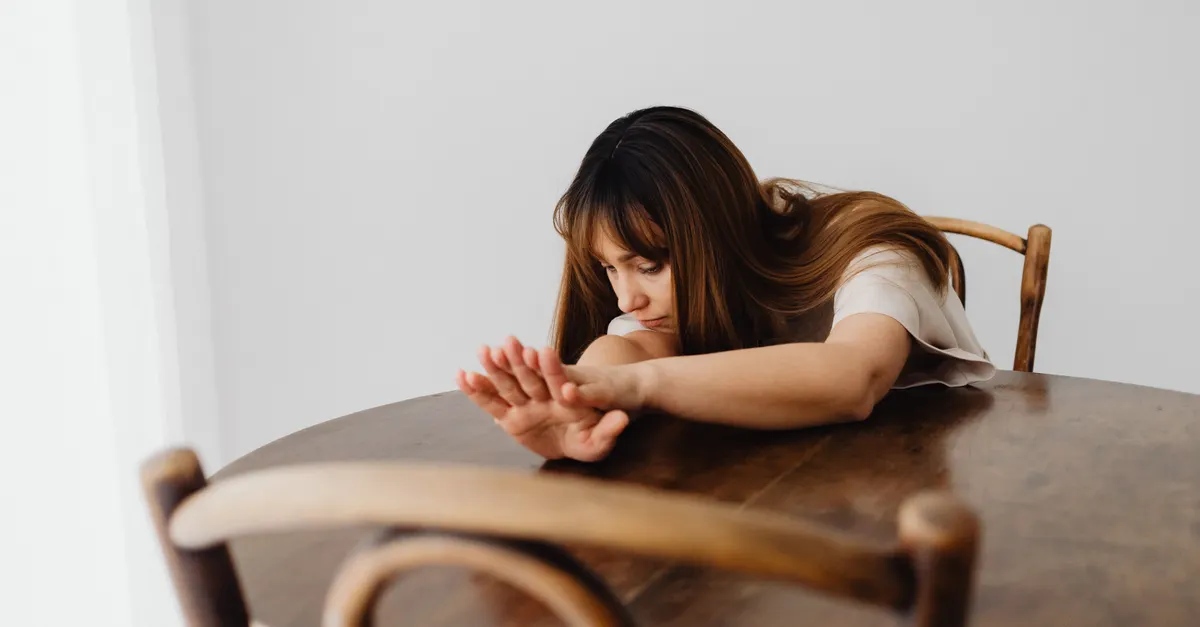 Woman sitting alone at a table with her arms resting on it, looking somber and contemplative.