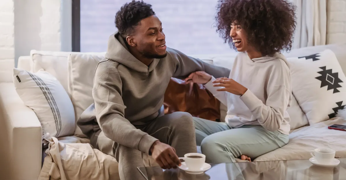 Positive African American couple talking and laughing on comfortable sofa at home in daytime.