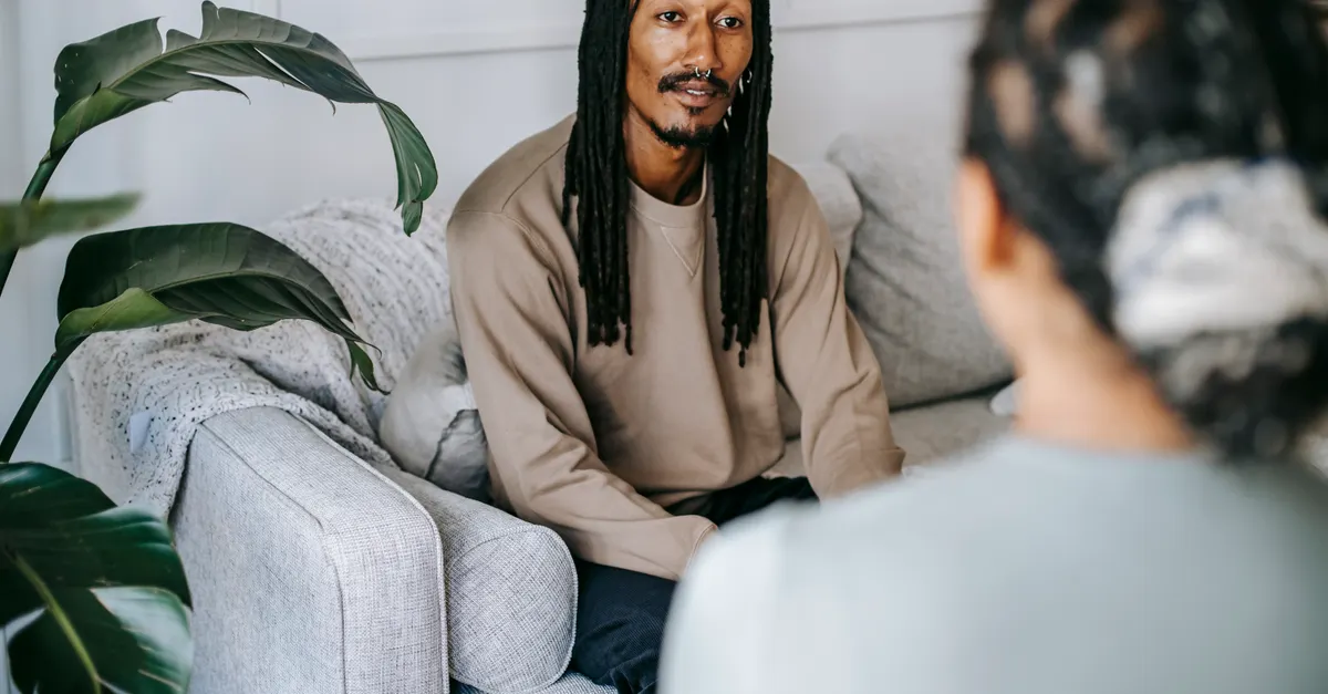 Man with dreadlocks sitting on a couch talking with a female friend in a cozy living room