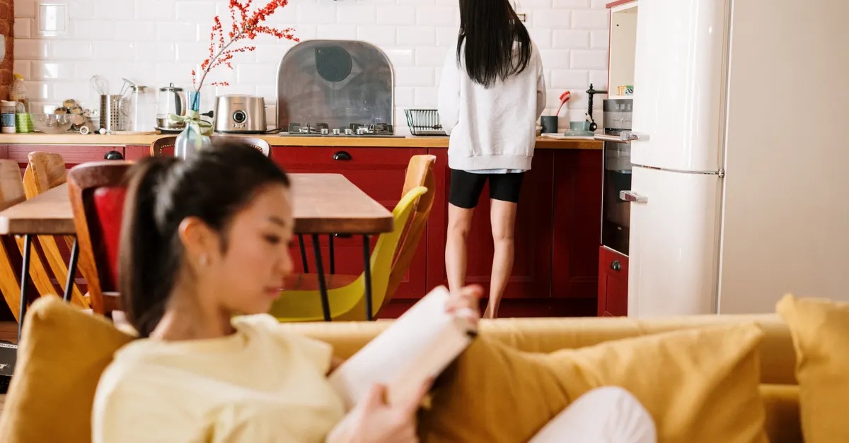 Two women at home, one reading on the sofa while the other stands at the kitchen counter