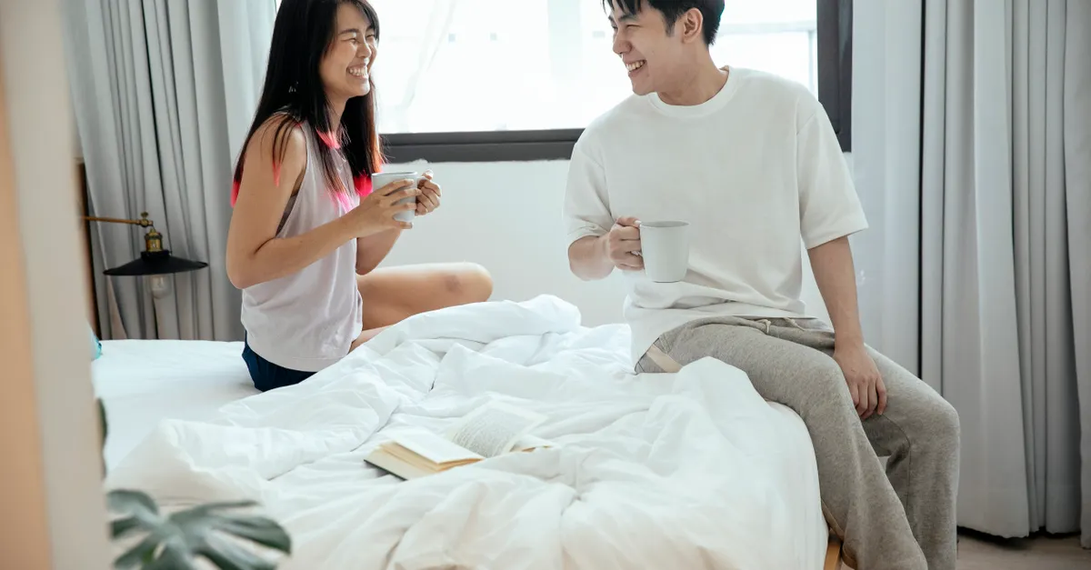 Asian couple sitting on bed, smiling at each other while holding coffee mugs.