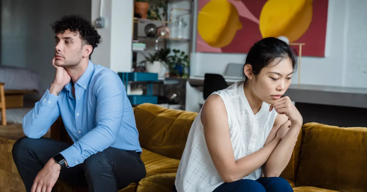 A couple sits indoors on a sofa, both appearing thoughtful and contemplative.