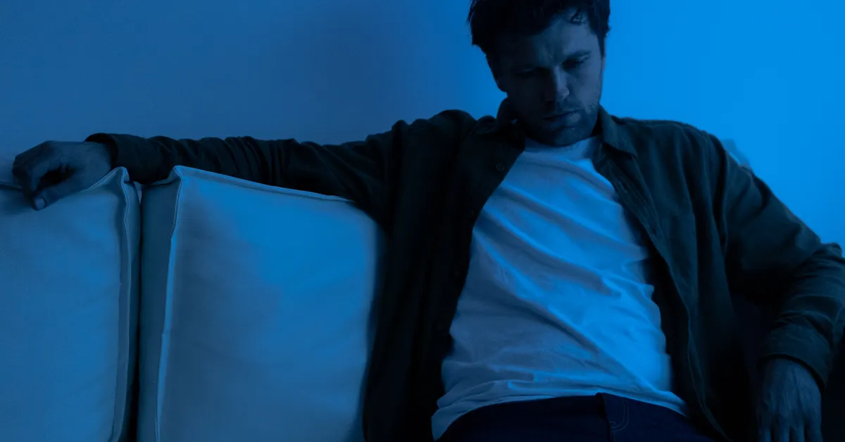 A man sits alone on a sofa under blue lighting, conveying a somber mood.